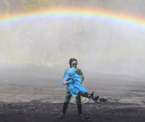 Photo of Gabriela Ciocarlie holding her child in rain ponchos in the rain with a rainbow